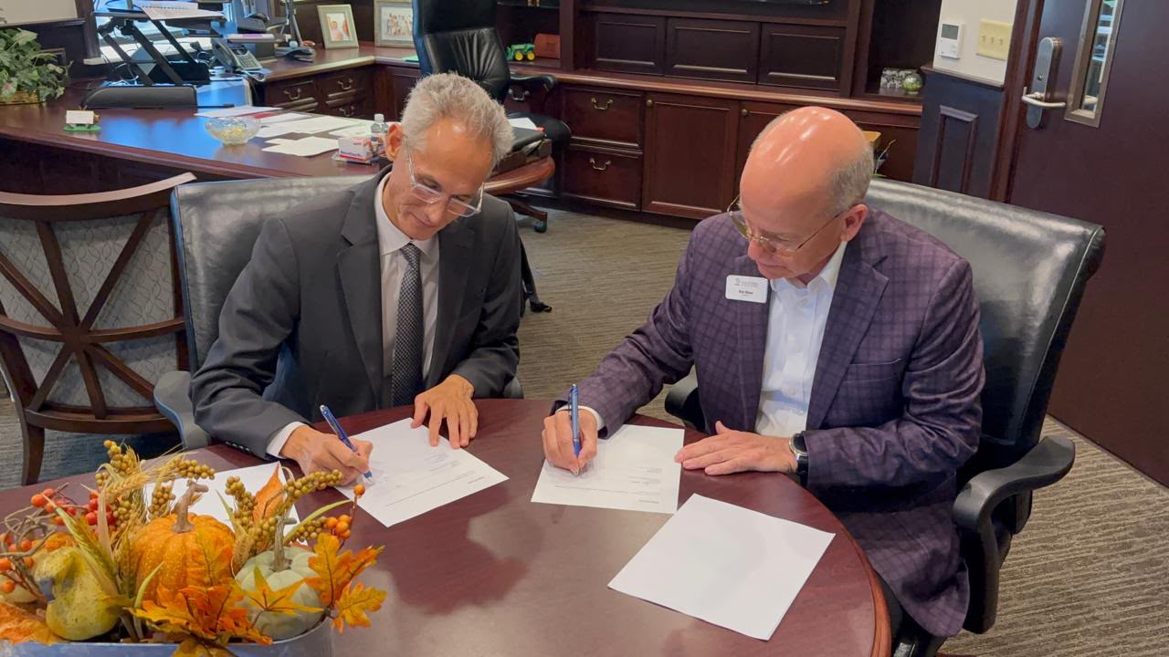 Two university representatives seated together as they sign documents at a conference table